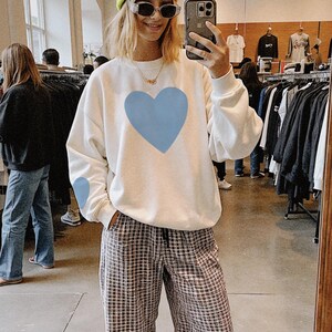 White crewneck sweatshirt featuring a large Carolina blue heart on the chest and a smaller matching Carolina blue heart on the right sleeve.