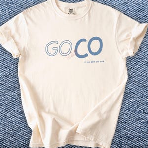 Puede incluir: Camiseta color crema con el texto azul "GO CO" y la frase "if you know you know" debajo. La camiseta tiene mangas cortas y cuello redondo.