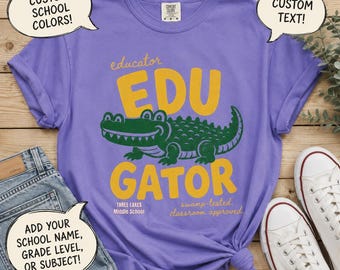 May include: A purple short-sleeve t-shirt featuring a green alligator graphic and yellow text that reads educator EDU GATOR, THREE LAKES MIDDLE SCHOOL, and swamp-tested, classroom approved. The shirt includes three speech bubbles with custom text.