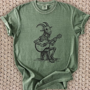 May include: Olive green t-shirt featuring a black illustration of a goat skeleton playing a guitar. The goat is seated on a tree stump, with the guitar labeled "Billy". The shirt has a crew neck and short sleeves.
