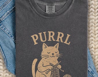 May include: Dark gray t-shirt with the word "PURRL" above a tan cat graphic knitting with needles and yarn. The cat is sitting next to a ball of yarn. The shirt is folded on top of blue jeans.