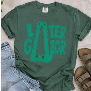 Blue Spruce Comfort Colors flat lay graphic tee featuring a playful Later Gator alligator illustration forming the letter A in chunky retro green typography.