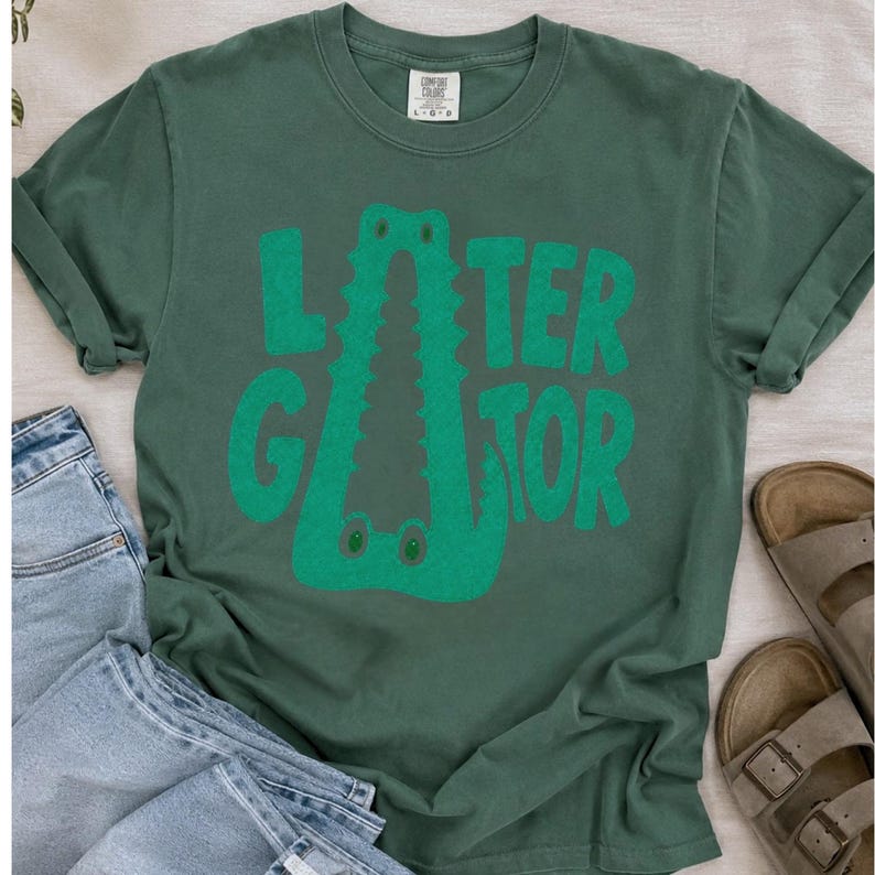 Blue Spruce Comfort Colors flat lay graphic tee featuring a playful Later Gator alligator illustration forming the letter A in chunky retro green typography.