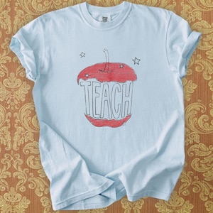 Light blue teacher shirt featuring hand drawn apple illustration and TEACH lettering, relaxed boho teacher tee with Free People inspired aesthetic