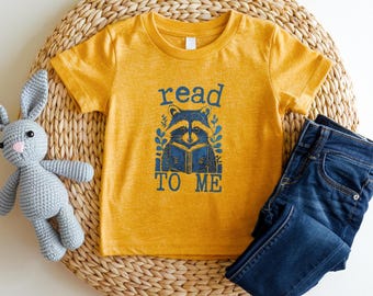 May include: Mustard yellow t-shirt with a blue graphic of a raccoon reading a book, with the words "read TO ME". The shirt is paired with dark blue jeans and a gray stuffed bunny on a woven mat.
