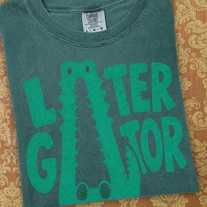 Blue Spruce Comfort Colors folded graphic tee featuring a playful Later Gator alligator illustration forming the letter A in chunky retro green typography.