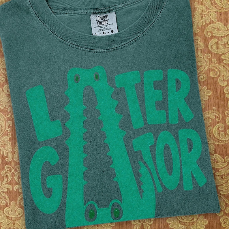 Blue Spruce Comfort Colors folded graphic tee featuring a playful Later Gator alligator illustration forming the letter A in chunky retro green typography.