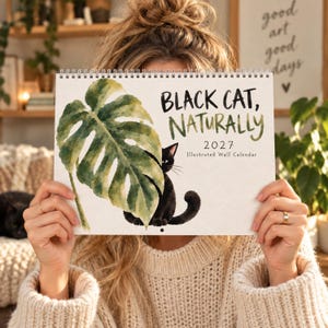 girl holding up 2027 black cat wall calendar with watercolor cat and green monstera leaf cover, illustrated calendar gift for cat lover