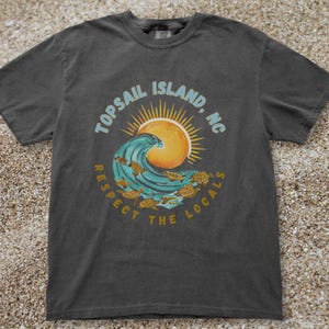 May include: A charcoal gray short-sleeve t-shirt featuring a graphic of a teal ocean wave, a yellow sun, and several sea turtles. The text reads Topsail Island, NC and Respect The Locals in a circular arrangement around the central design.