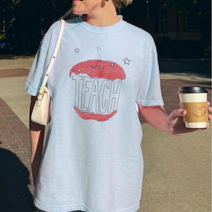 Light blue teacher shirt featuring hand drawn apple illustration and TEACH lettering, relaxed boho teacher tee with Free People inspired aesthetic