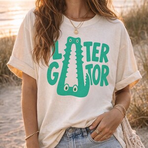 IVORY Comfort Colors vacation graphic tee featuring a playful Later Gator alligator illustration forming the letter A in chunky retro green typography.