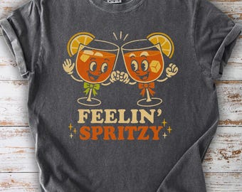 May include: Dark gray t-shirt featuring a retro design with two cartoon cocktail glasses clinking. The glasses have orange liquid, ice cubes, and lemon slices. The text "FEELIN' SPRITZY" is below the glasses, with star accents.