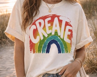 May include: An off-white short-sleeve t-shirt featuring a graphic of a rainbow with a distressed, painted texture. The word CREATE is printed in large, bold, white capital letters across the center of the rainbow design on the front of the shirt.