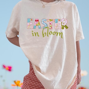 May include: Cream-colored t-shirt with the text "EASTER in bloom" in a patchwork style. The word "EASTER" is in pastel colors, and "in bloom" is in green. The shirt is worn with a red and white checkered skirt.