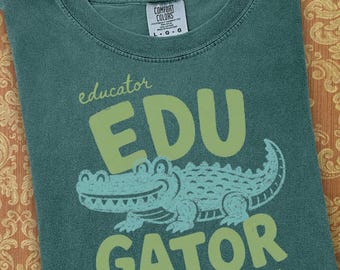 May include: A teal crew neck t-shirt featuring a light green graphic of an alligator. The text reads "educator EDU GATOR" and "swamp-tested. classroom approved." The shirt has a Comfort Colors brand label at the neck indicating a size large.