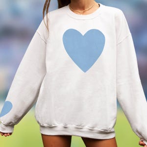 White crewneck sweatshirt featuring a large Carolina blue heart on the chest and a smaller matching Carolina blue heart on the right sleeve.