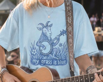 May include: A light blue short-sleeve t-shirt featuring a dark blue graphic of a goat playing a banjo in a field of grass. The text "my grass is blue" is printed in dark blue below the goat. The shirt has a crew neckline and a relaxed fit.