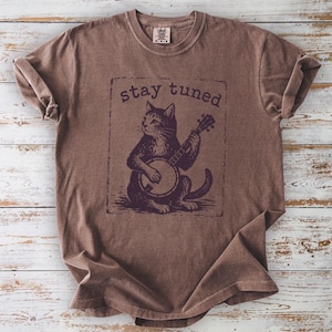 May include: A brown short-sleeve t-shirt featuring a dark brown graphic of a cat playing a banjo inside a rectangular border. The text "stay tuned" is printed in a dark brown font above the cat illustration. The shirt has a crew neckline.