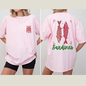Distressed oversized graphic tee with a vintage fisherman aesthetic. Small red sardines graphic on the front chest, full-size distressed red sardines print on the back with sardines in green script. Nautical, retro inspired design with a worn-in look