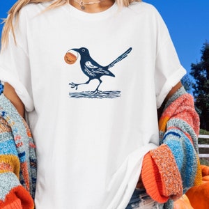 May include: A white t-shirt with a black and white illustration of a bird carrying a small pumpkin. The bird is perched on a branch.
