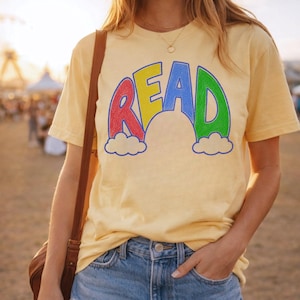 May include: A light yellow t-shirt with the word "READ" in a rainbow design. The letters are red, yellow, blue, and green, with white clouds at the bottom. The shirt is paired with blue denim jeans.