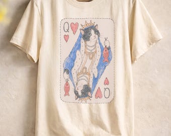 May include: A cream-colored short-sleeve t-shirt featuring a graphic print of a playing card design. The illustration depicts a black and white cat wearing a crown and blue robes, holding a fish, with the letter Q and heart symbols in the corners.