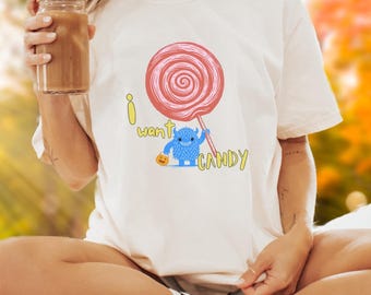 May include: Cream-colored t-shirt featuring a large red and pink lollipop design. A blue monster with a pumpkin bucket is holding the lollipop, with the words "I want CANDY" in yellow. The shirt is a casual, everyday wear item.