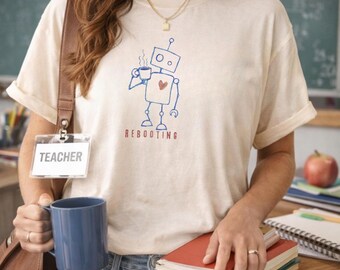 May include: A cream-colored short-sleeve t-shirt featuring a blue line-art graphic of a robot holding a steaming mug with a small red heart on its chest. Below the robot, the word REBOOTING is printed in red capital letters on the front of the shirt.