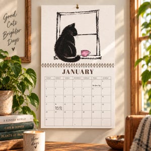 2027 black cat calendar hanging open to January with whimsical watercolor cat by a window and pink coffee cup