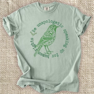 May include: A light green t-shirt with a green bird graphic inside a circle that reads "Be unapologetic speaking up for human rights." The bird has intricate floral and leaf details. The shirt is folded.