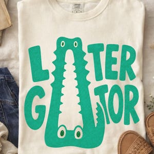 IVORY Comfort Colors folded graphic tee featuring a playful Later Gator alligator illustration forming the letter A in chunky retro green typography.