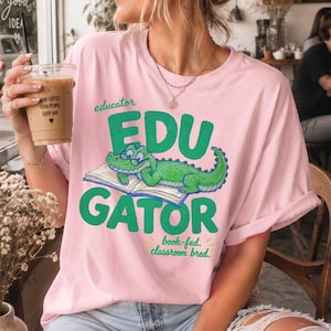 May include: A light pink short-sleeve t-shirt featuring a green cartoon alligator wearing glasses and lying on an open book. The text reads "educator EDU GATOR" and "book-fed. classroom bred." in green lettering across the front of the garment.