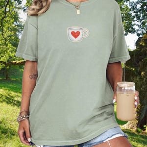 May include: Sage green t-shirt featuring a stitched design of a coffee cup with a red heart. The shirt has short sleeves and a relaxed fit. The model is wearing denim shorts and holding a glass of iced coffee.
