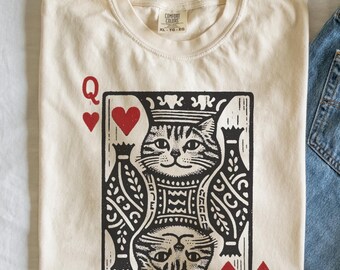 May include: A cream colored t-shirt with a black and white illustration of the Queen of Hearts playing card featuring a cat. The card has red hearts in the corners and the letter Q in the top left corner.