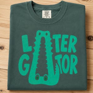 Blue Spruce Comfort Colors folded graphic tee featuring a playful Later Gator alligator illustration forming the letter A in chunky retro green typography.