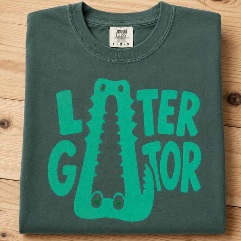 Blue Spruce Comfort Colors folded graphic tee featuring a playful Later Gator alligator illustration forming the letter A in chunky retro green typography.