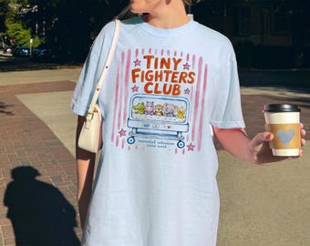 May include: A light blue t-shirt featuring a graphic of a hospital crib containing five small cartoon animals. The text reads "TINY FIGHTERS CLUB" and "neonatal intensive care unit" in brown and blue, surrounded by pink vertical stripes and stars.