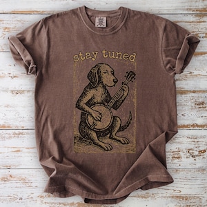 May include: Brown t-shirt featuring a dog playing a banjo. The dog illustration is in a vintage style, with the words "stay tuned" above it. The shirt has a crew neck and short sleeves, and is made of a soft, comfortable material.