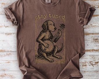 May include: Brown t-shirt featuring a dog playing a banjo. The dog illustration is in a vintage style, with the words "stay tuned" above it. The shirt has a crew neck and short sleeves, and is made of a soft, comfortable material.
