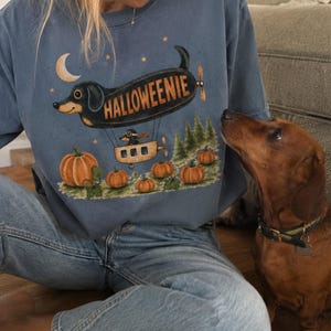 May include: A blue crewneck sweatshirt featuring a graphic print of a black dachshund shaped like a blimp with the text HALLOWEENIE. The design includes a small pilot dachshund in a basket, orange pumpkins, green pine trees, and a crescent moon.