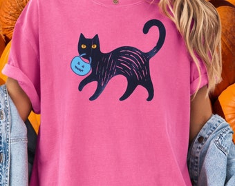 May include: A pink t-shirt featuring a black cat carrying a blue pumpkin-shaped candy bag. The cat has yellow eyes and a curved tail. The shirt is a solid pink color, perfect for Halloween.