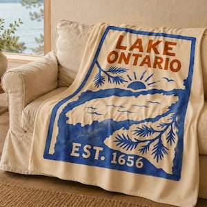 May include: A cream-colored throw blanket featuring a large blue rectangular graphic with the text LAKE ONTARIO in orange, a stylized sun, pine branches, and the text EST. 1656 in white. The design is printed on a soft, textured fabric surface.