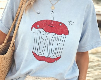 May include: A light blue short-sleeve t-shirt featuring a red apple graphic with the word TEACH printed in large black letters across the center. The design includes small black star outlines surrounding the apple. The shirt has a crew neckline.