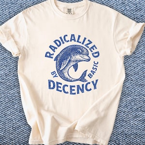 May include: Cream-coloured t-shirt featuring a blue dolphin graphic and the words "RADICALIZED BY DECENCY BASIC". The shirt has short sleeves and a crew neck. The material looks soft and comfortable.
