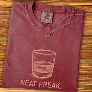 以下が含まれることがあります： リキュールのグラスのスケッチと「NEAT FREAK」の文字が描かれた、マルーン色のComfort Colors Tシャツ。小さなペンダントが付いたシルバーのネックレスがシャツの上にあります。シャツは木製の表面にあります。