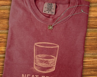 May include: A maroon Comfort Colors t-shirt with a sketched image of a glass of liquor and the words "NEAT FREAK." A silver necklace with a small pendant rests on the shirt. The shirt is on a wooden surface.
