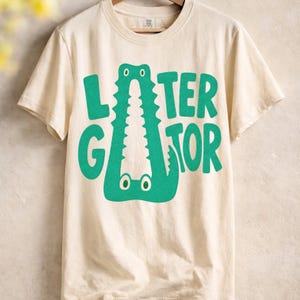 Hanging IVORY Comfort Colors  graphic tee featuring a playful Later Gator alligator illustration forming the letter A in chunky retro green typography.