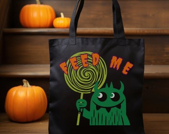 May include: Black tote bag with a green monster graphic holding a green and yellow lollipop. The text "FEED ME" is in orange and purple. Orange pumpkins are in the background.