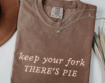May include: A brown Comfort Colors t-shirt with the text "keep your fork THERE'S PIE" in a light brown color. A silver necklace with a small pendant is draped across the shirt. The shirt is folded on a white surface.