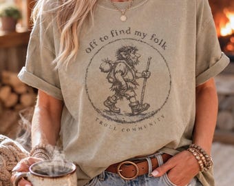 May include: Olive green t-shirt with a circular graphic of a troll carrying a pack and walking with a staff. The text "off to find my folk" and "Troll Community" are printed around the graphic. The shirt has rolled-up sleeves.